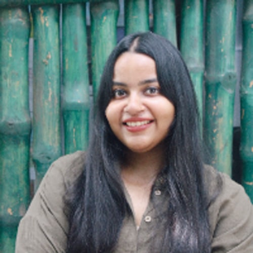 A smiling woman with long black hair poses in front of a green bamboo backdrop, wearing a casual olive shirt.